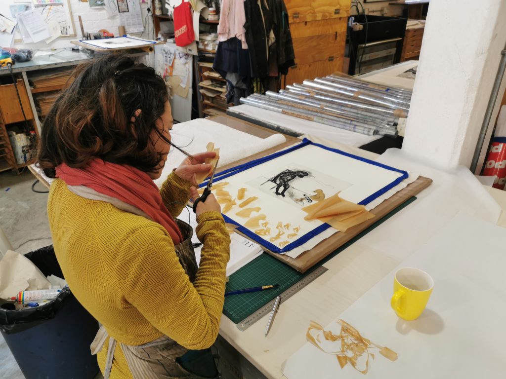 Editioning printer, Kim-Lee Loggenberg, standing at a table in front of an edition of Wilma Cruise’s print titled Word. She is cutting out tea-stained chine-collé pieces to be adhered to the print.