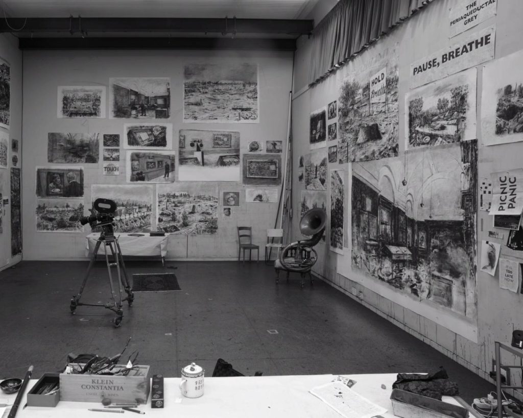 Black and white photograph of William Kentridge’s studio. The walls are covered with black and white charcoal drawings, mainly still life, hung salon style from floor to ceiling. A work table sits in the foreground with art supplies on it. A camera on a wheeled tripod sits in the center of the room. A tuba and two chairs sit in the back corner.