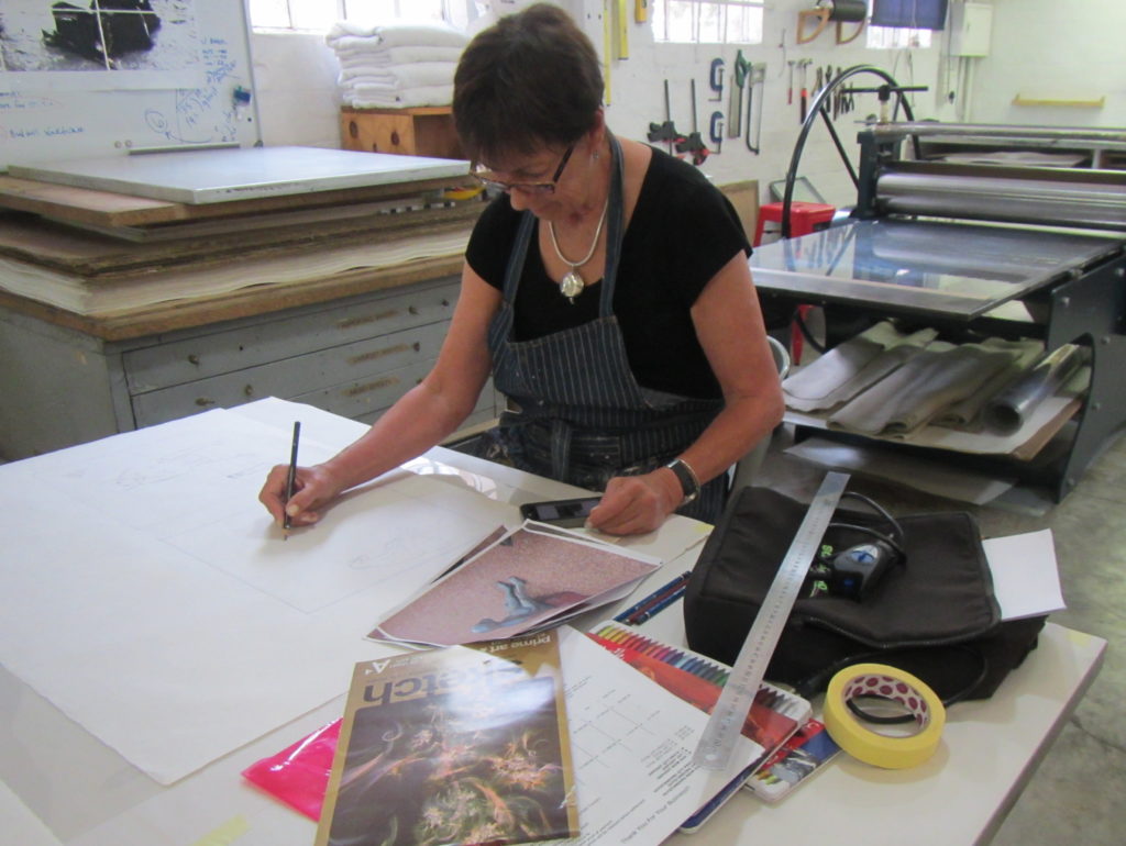 Artist Wilma Cruise sitting at a table surrounded by reference photos and art supplies sketching a baboon on a large white piece of paper to feature in her print titled The End Game.