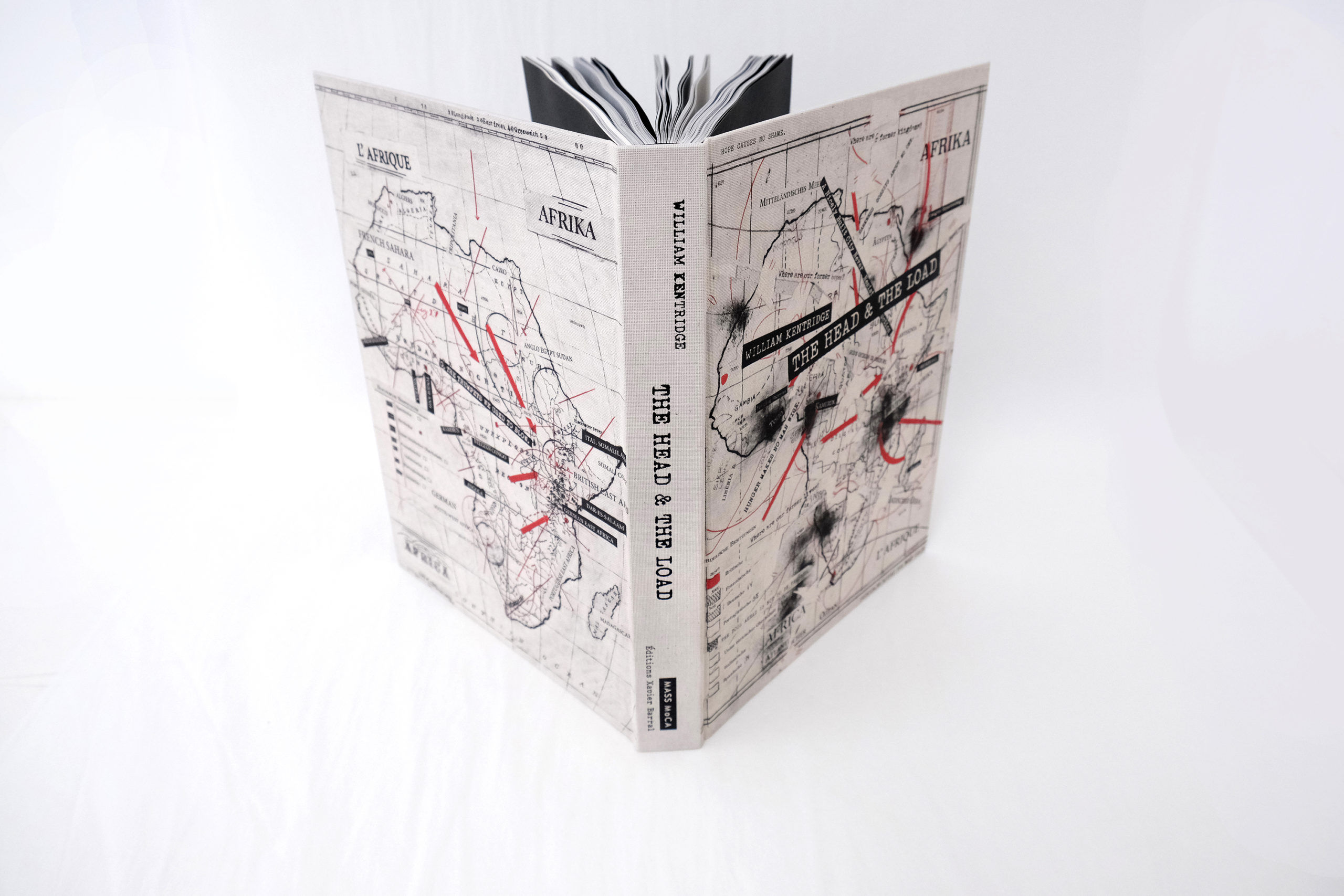 William Kentridge's The Head and the Load book standing up on its spine, open with the cover facing the camera. The cover art is a black and white illustrated map of Africa with black text labels and red arrows pointing to areas on the map. The text “The Head and the Load” and “William Kentridge” appears on the spine and the front cover.