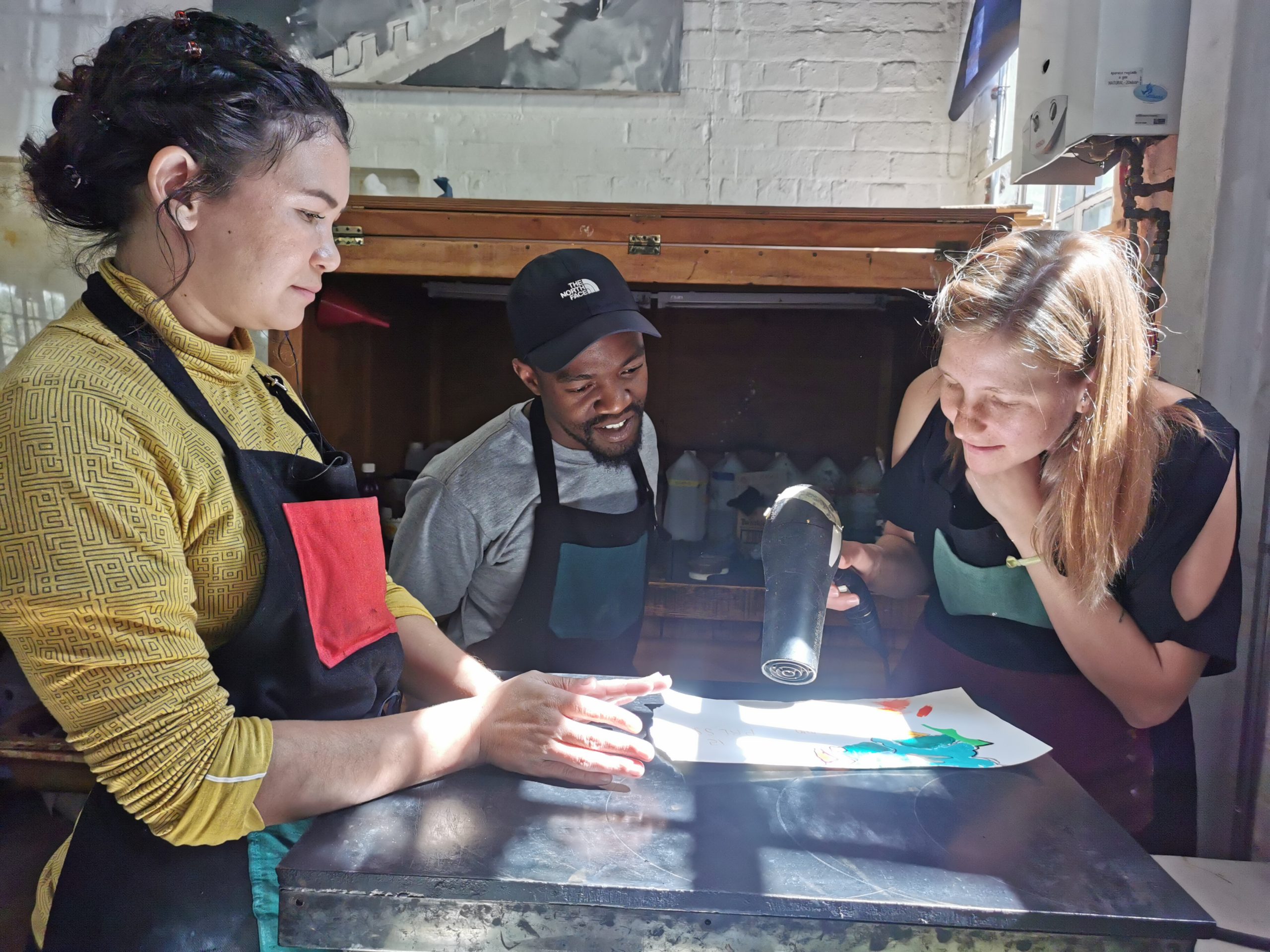 Sarah Judge (right) uses a heat gun to activate 'puff paint' to be collaged on Khotso Motsoneng's (centre) works; right: Kim-lee Loggenberg